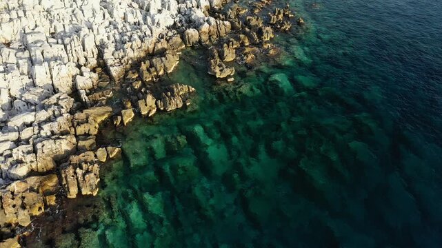 Aerial view of rugged rocky coastline meeting crystal clear turquoise waters at Cap Tenare. The scene highlights the natural beauty and dramatic textures of the coastal landscape.