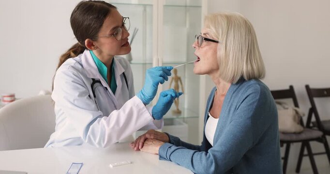 Female doctor wearing gloves performs oral swab test, collect sample from middle age woman patient mouth during medical checkup. Clinic procedures, diagnostics analysis, testing of viral infections