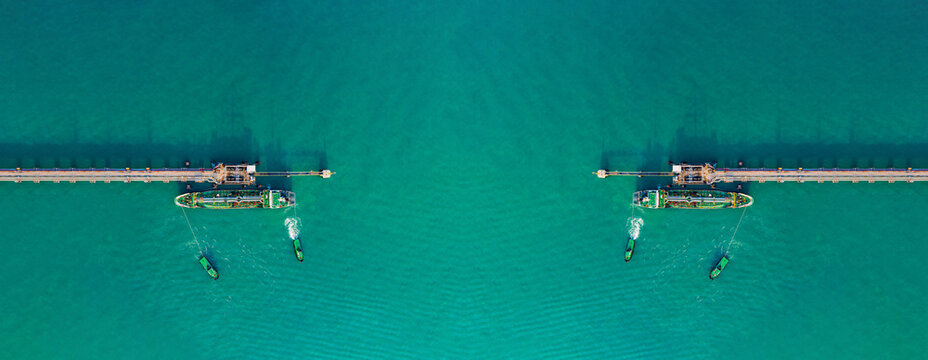 Aerial top view drone shot of industrial oil tanker docking at offshore pipeline terminal in turquoise ocean water during daylight. Long pier structure connects energy infrastructure.