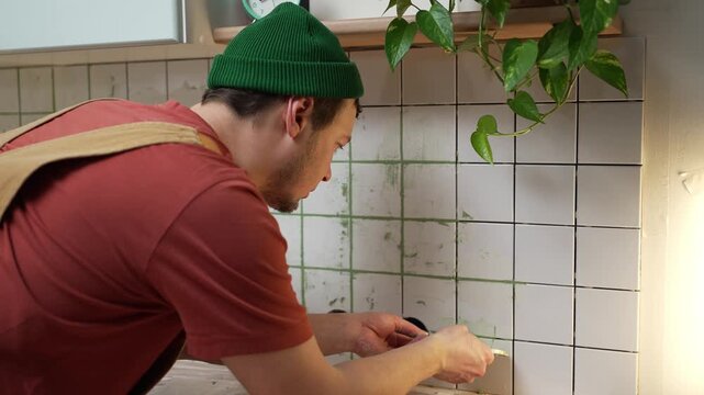 Young handyman in overalls carefully applying green grout between white ceramic tiles on a kitchen backsplash, meticulously finishing the seams with a special tool during a home renovation project