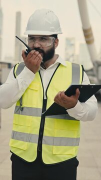 Portrait of bearded Indian contractor with clipboard talking on portable radio at construction site. Skilled manager discusses building plans against bridge columns vertical shot