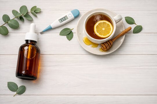 Top view of hot tea with lemon, honey, digital thermometer, eucalyptus leaves and medicine bottle on white wooden background
