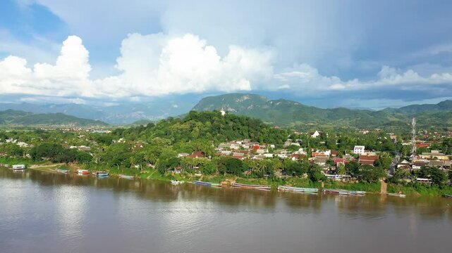 Scenic aerial shot of Luang Prabang with lush greenery, riverside boats, and surrounding mountains under a dramatic sky. The tranquil riverfront cityscape highlights the natural beauty of Laos.