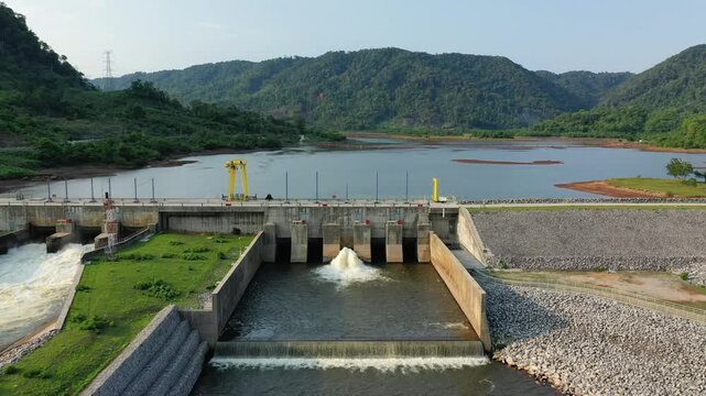 A modern hydroelectric dam set amidst lush green hills and a reservoir in the Laotian countryside between Phonsavan and Phou Khoun. The scene features clear water, engineered structures, and natural