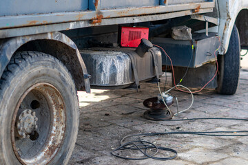 Fototapeta premium Charging a truck battery with a red charger on a construction site, featuring makeshift electrical connections, cable reels, and vehicle chassis details, documenting technical aspects of fleet mainten