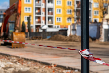 Close-up of red and white warning tape tied to a post, securing a construction site with an excavator and yellow apartment block in blurred background, emphasizing safety during urban investments. © Adam