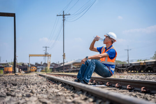 Railway worker sitting on tracks drinking water to stay hydrated under hot sun, Tired technician taking a break at railway depot, Worker man hydrating during a break on railroad maintenance site