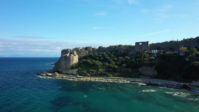 Aerial view of a historic stone fortress overlooking the turquoise sea and lush greenery in Koroni, Greece. Clear blue sky and coastal landscape create a serene Mediterranean scene.