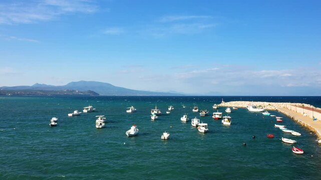 Small fishing boats anchored in the clear blue waters of Koroni harbor, with mountains and a stone pier in the background under a bright sky.
