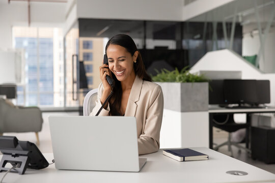 Smiling New Zealander professional business woman talking on landline phone at workplace desk using laptop, discuss details of contract or agreement with corporate client, provide services remotely