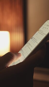 Hand holding smartphone and scrolling social media feed in dark room with desk on background, illuminated by warm tone light. Digital addiction and mindless content consuming