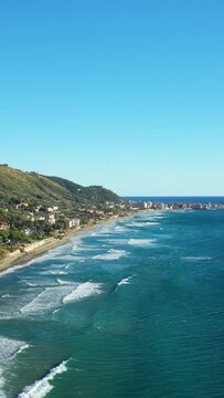 Scenic aerial view of the Acciaroli coastline with turquoise waves, sandy beach, and lush hills under a clear blue sky in southern Italy.