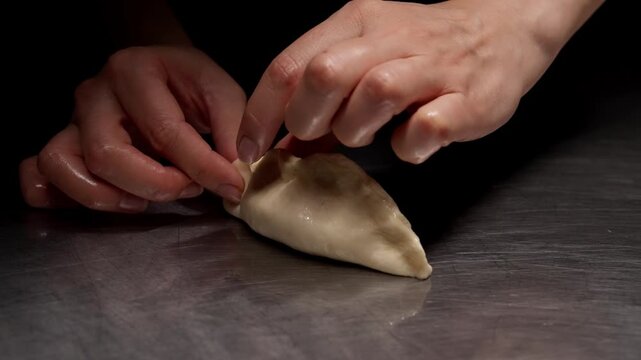 Close-up sequence of skilled hands shaping and sealing a stuffed dumpling on a reflective metal surface, highlighting delicate finger movements and handcrafted culinary detail