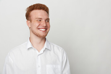Smiling young man with red hair wearing white shirt looks away on light background. Casual portrait...