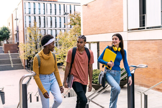 Group of diverse college students walking up university campus stairs, carrying books and backpacks, discussing while moving