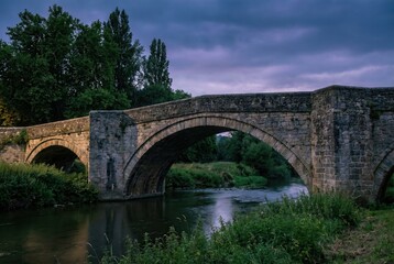 Fototapeta premium Old stone arch bridge over river at dusk with dramatic cloudy sky 