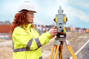 Young female surveyor operates a total station at a construction site while wearing safety hi-viz jacket
