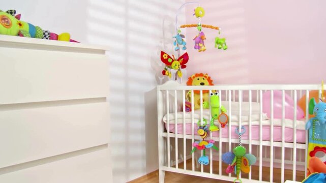 Cozy nursery room interior with white wooden crib and chest of drawers featuring colorful hanging plush toys and baby mobile for newborn child bedroom design