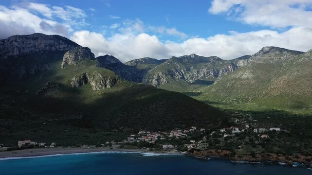 Aerial view of a small coastal village nestled between lush green hills and dramatic mountains near Monemvasia, Greece. The turquoise sea contrasts beautifully with the rugged landscape under a partly