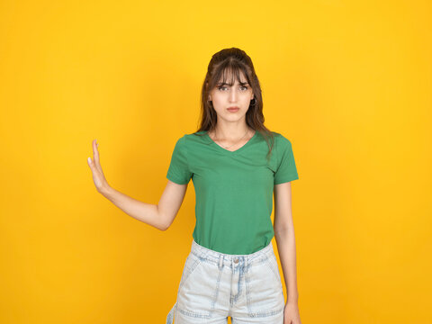 Serious young woman in green t-shirt making a stop gesture with her hand aside against a yellow background. Concept of refusal, setting boundaries, or warning.