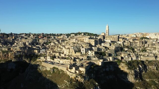 Aerial view of the historic stone buildings and dramatic cliffs of Matera, Italy, under a clear blue sky. The iconic old town and cathedral tower are visible in this panoramic landscape.