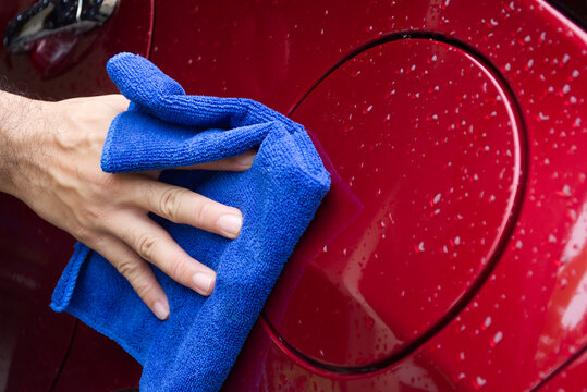 Car Paint Surface Detailing, A hand uses a blue microfiber cloth to dry and polish the wet, red paint surface of a car door, focusing on the fuel filler flap.