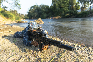 Obraz premium Focused soldier in camouflage aiming rifle while lying prone on riverbank. This military special force member in jungle on combat mission remains vigilant outdoors