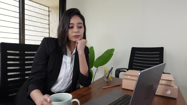 Close up of thoughtful young Indian businesswoman boss manager sits on chair at workplace desk look at laptop thinking pondering planning with serious face expression.