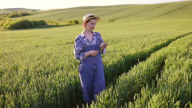 A young woman wearing overalls and a straw hat stands in a lush green wheat field, holding a stalk of wheat and looking thoughtfully into the distance
