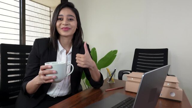 Closeup portrait of happy confident beautiful young Asian business woman boss manager sits on chair at workplace desk looking at camera show thumb up hand gesture agree approve ok like sign.