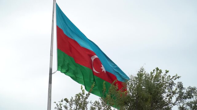 The Azerbaijani flag waves proudly in the wind, set against a backdrop of trees and a cloudy sky, symbolizing national pride and identity.