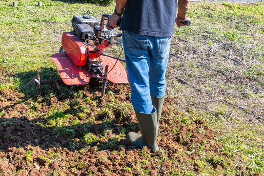 Farmer in rubber boots is tilling soil with a motor cultivator, preparing the ground for planting, emphasizing sustainable agriculture and rural lifestyle