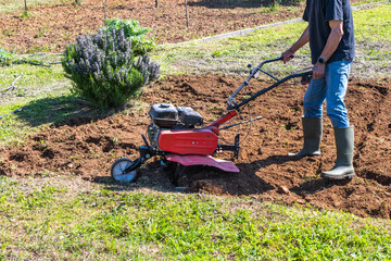 Person operating a small red rototiller, turning brown soil in a sunny outdoor garden, preparing the ground for planting crops and cultivating a homestead for self-sufficiency