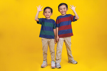 Fototapeta premium Happy and Smiling Asian Boy Stands and Waves His Hand on Yellow Background