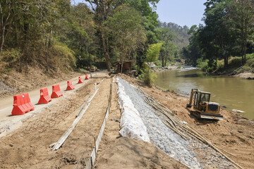 Naklejka premium Construction site for road development and riverbank engineering. An excavator works on dirt infrastructure project next to calm river, showing progress and industry