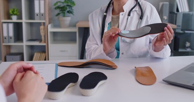 Orthopedic doctor sits at workplace desk holding insoles, explaining benefits, recommending foot support solution to woman patient, close-up. Flat foot correction, musculoskeletal health concept