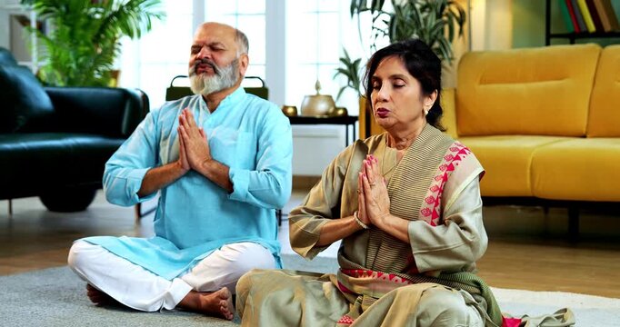 Indian senior couple doing omkar pranayama or chanting mantra or yoga at home in the morning sitting with folded legs in modern living room, practicing mindful breathing for health