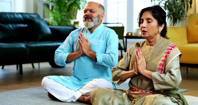 Indian senior couple doing omkar pranayama or chanting mantra or yoga at home in the morning sitting with folded legs in modern living room, practicing mindful breathing for health