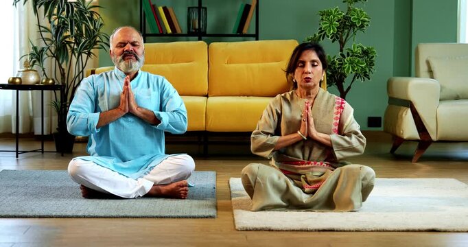 Indian senior couple doing omkar pranayama or chanting mantra or yoga at home in the morning sitting with folded legs in modern living room, practicing mindful breathing for health