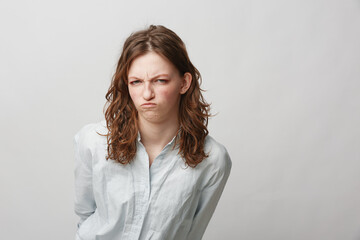 Obraz premium Young woman with curly hair wearing white shirt shows displeased facial expression, frowning and looking at camera. Isolated on light grey background. Concept of dissatisfaction and emotion.