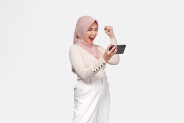 Happy successful Asian Muslim woman in hijab holding smartphone with fist raised.