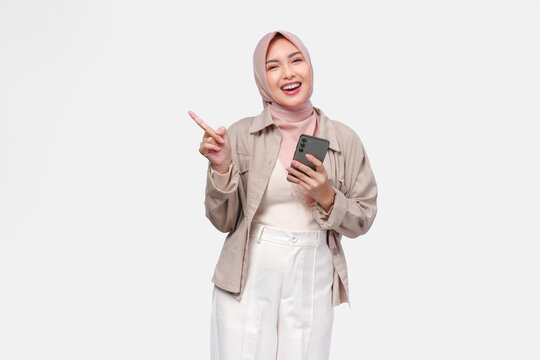 Pretty young Muslim woman holding a smart phone while pointing to the copy space on her side.