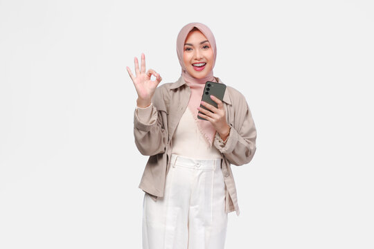 Joyful young Asian woman in hijab holding a smartphone while showing okay finger gesture.