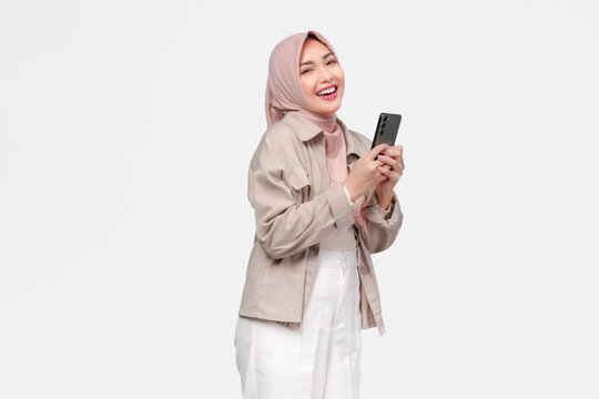 Excited young Asian Muslim woman in hijab holding smartphone with happy expression.