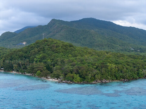 Aerial view of the lush green island meeting the turquoise sea under a cloudy sky, Tanjung Gelam Beach, Karimunjawa, Central Java, Indonesia.