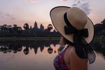 Woman wearing straw hat observing the iconic angkor wat temple at sunrise with water reflection
