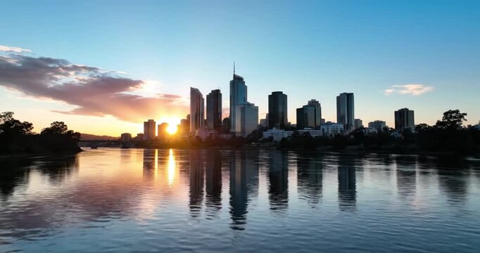 An impressive urban skyline dramatically silhouettes against a vivid sunset or sunrise, reflecting golden and warm hues across the tranquil river below. Modern skyscrapers stand tall, creating a majes