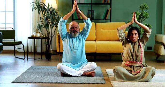 Indian senior couple doing omkar pranayama or yoga at home in the morning sitting with folded legs in modern living room chanting aum, practicing mindful breathing exercise for health