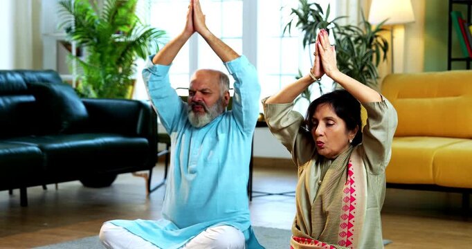 Indian senior couple doing omkar pranayama or yoga at home in the morning sitting with folded legs in modern living room chanting aum, practicing mindful breathing exercise for health