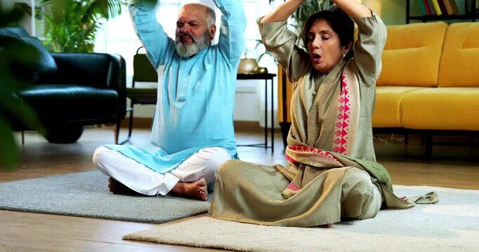 Indian senior couple doing omkar pranayama or yoga at home in the morning sitting with folded legs in modern living room chanting aum, practicing mindful breathing exercise for health